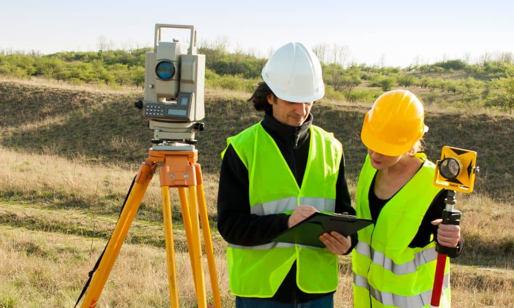 Two land surveyors using surveying equipment on sloped terrain, collecting elevation and drainage data before construction on uneven land in Pennsylvania.