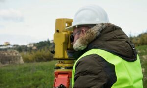 Licensed land surveyor conducting a property survey using surveying equipment outdoors while wearing safety gear